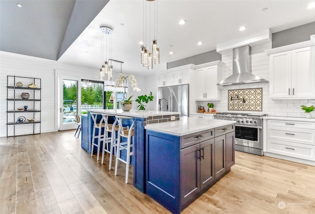 kitchen with white cabinetry, stainless steel appliances, an island with sink, hanging light fixtures, and wall chimney range hood