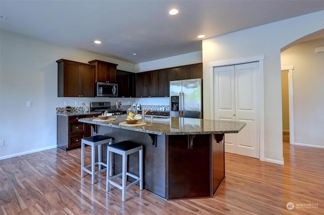 kitchen with light wood-type flooring, appliances with stainless steel finishes, stone countertops, a kitchen bar, and a kitchen island with sink