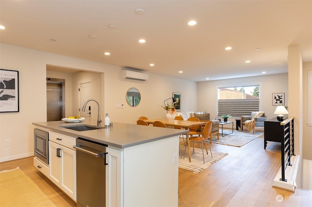 kitchen featuring an island with sink, sink, a wall mounted AC, white cabinetry, and light wood-type flooring