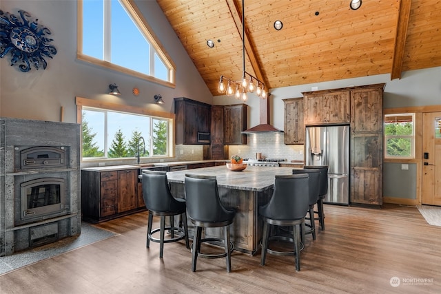 kitchen with a kitchen island, wall chimney range hood, light wood-type flooring, high vaulted ceiling, and stainless steel refrigerator with ice dispenser