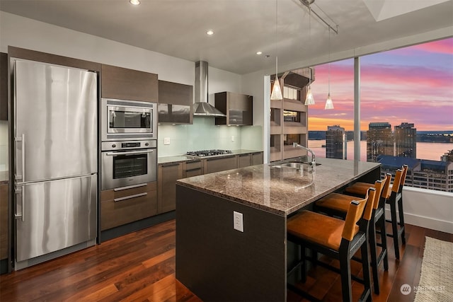 kitchen with sink, a breakfast bar area, a kitchen island with sink, stainless steel appliances, and wall chimney range hood