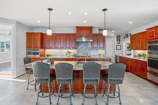 kitchen featuring light stone counters, double oven, decorative light fixtures, and an island with sink