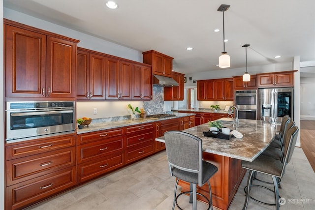 kitchen with decorative light fixtures, a kitchen bar, stainless steel appliances, light stone countertops, and a center island with sink