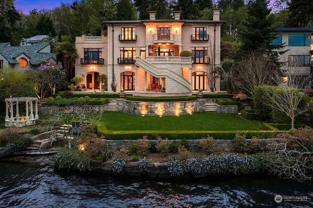 back house at dusk featuring a lawn, a water view, and a balcony