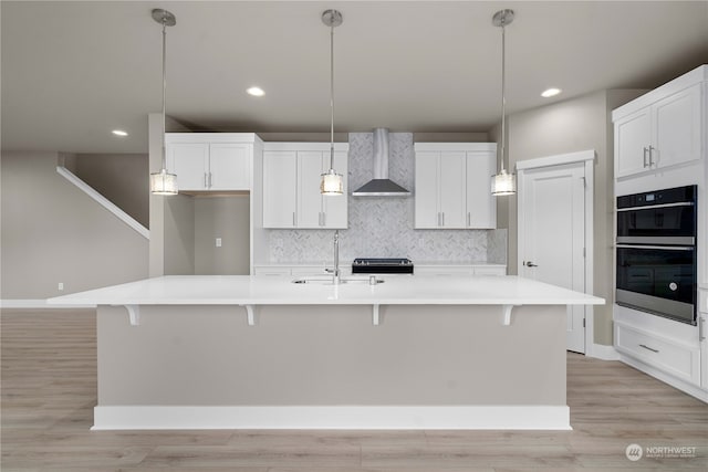 kitchen featuring a kitchen island with sink, light wood-type flooring, wall chimney exhaust hood, and double oven