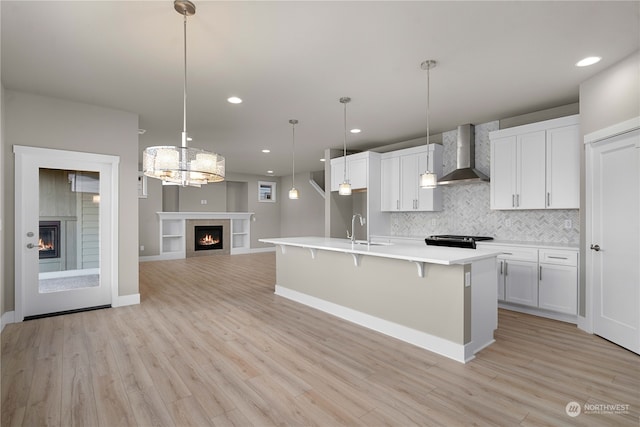 kitchen featuring sink, an island with sink, wall chimney range hood, white cabinetry, and light wood-type flooring