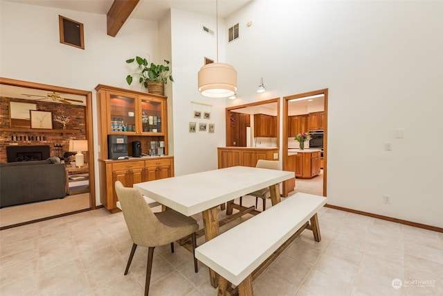 dining room featuring a ceiling fan, a brick fireplace, baseboards, beamed ceiling, and a towering ceiling