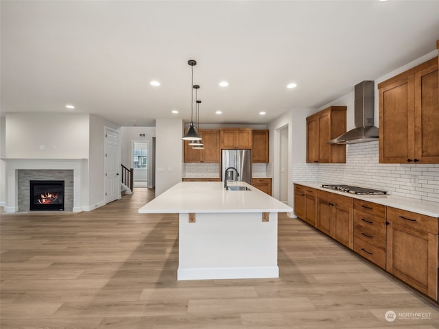 kitchen with wall chimney range hood, hanging light fixtures, light wood-type flooring, and an island with sink