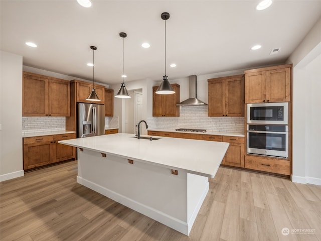 kitchen with sink, hanging light fixtures, stainless steel appliances, wall chimney exhaust hood, and a kitchen island with sink