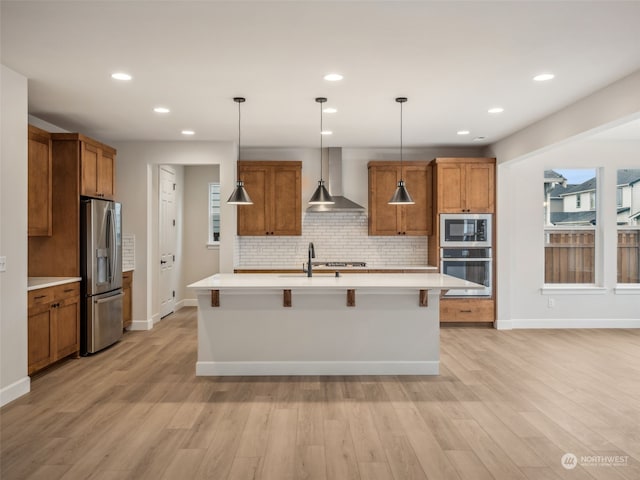 kitchen with wall chimney exhaust hood, appliances with stainless steel finishes, light hardwood / wood-style flooring, and an island with sink