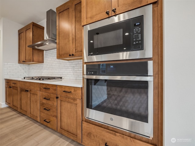 kitchen featuring stainless steel appliances, wall chimney range hood, light hardwood / wood-style flooring, and tasteful backsplash