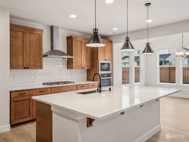 kitchen featuring a kitchen island with sink, wall chimney exhaust hood, sink, decorative light fixtures, and light hardwood / wood-style floors