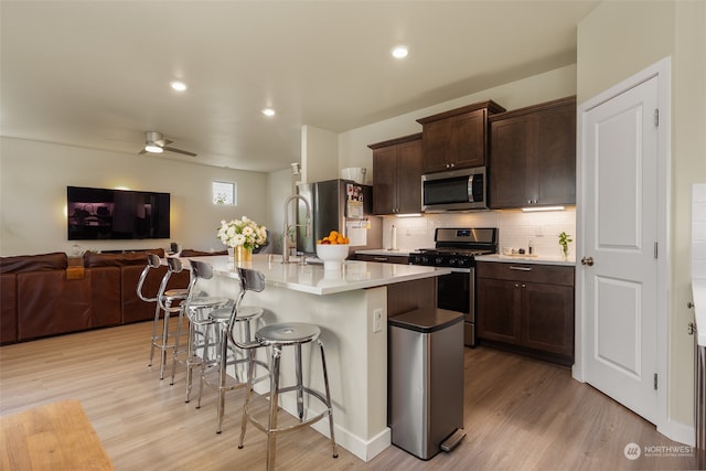 kitchen with appliances with stainless steel finishes, light wood-type flooring, a kitchen bar, ceiling fan, and a center island with sink