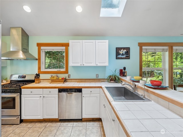 kitchen featuring appliances with stainless steel finishes, wall chimney exhaust hood, a healthy amount of sunlight, tile counters, and sink