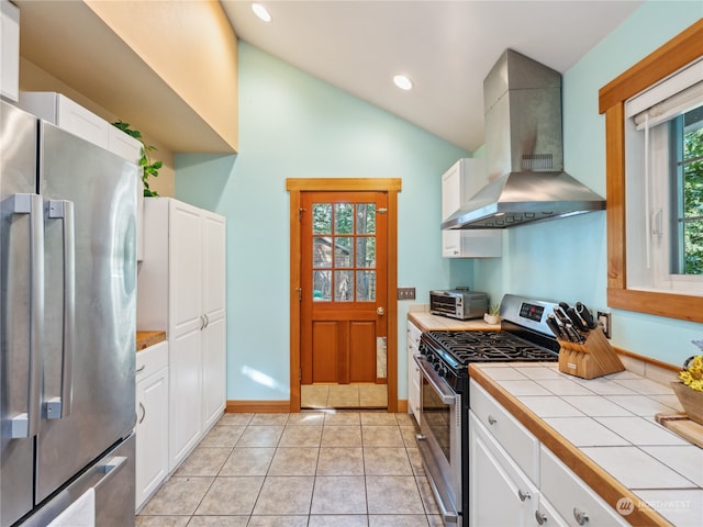 kitchen with appliances with stainless steel finishes, white cabinets, light tile patterned floors, tile counters, and wall chimney range hood