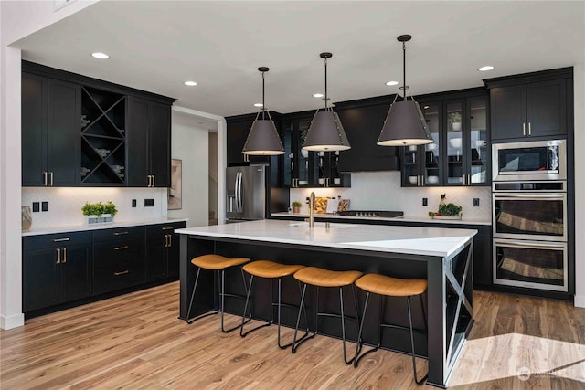 kitchen with decorative light fixtures, a kitchen island with sink, stainless steel appliances, and light hardwood / wood-style flooring