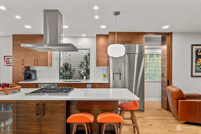 kitchen featuring hanging light fixtures, island exhaust hood, stainless steel appliances, light wood-type flooring, and a kitchen bar