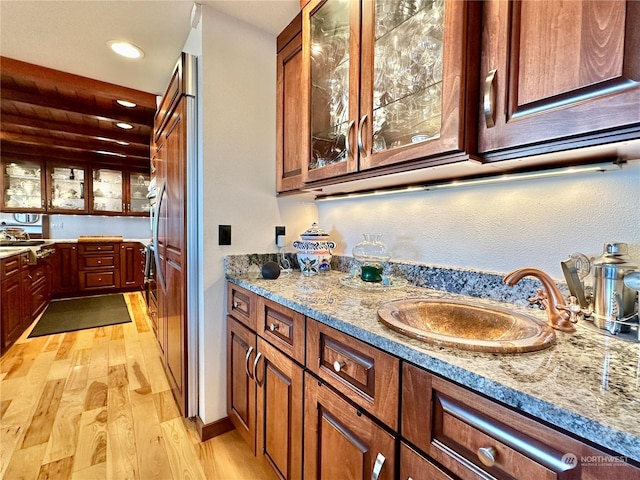 kitchen with glass insert cabinets, light stone countertops, light wood-style floors, brown cabinets, and a sink