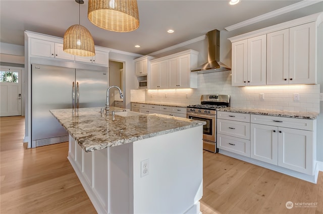 kitchen featuring white cabinets, an island with sink, hanging light fixtures, wall chimney range hood, and appliances with stainless steel finishes