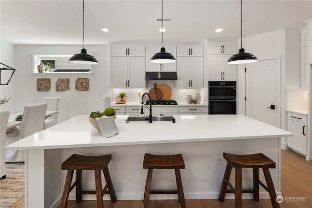 kitchen with hanging light fixtures, a kitchen island with sink, light hardwood / wood-style flooring, white cabinetry, and black double oven