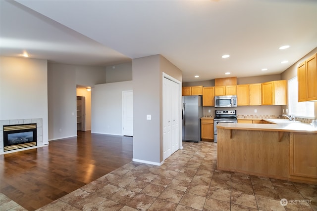 kitchen with kitchen peninsula, a tiled fireplace, stainless steel appliances, light brown cabinetry, and hardwood / wood-style floors