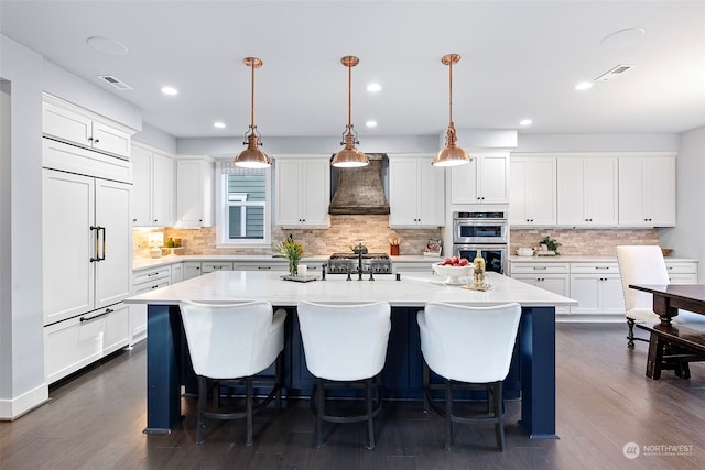 kitchen with custom range hood, hanging light fixtures, a kitchen island with sink, and dark wood-type flooring