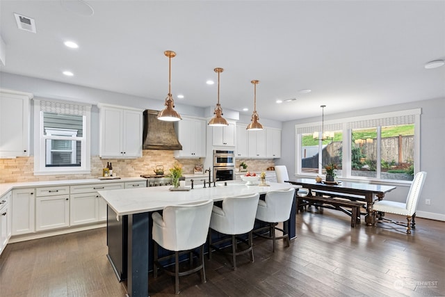 kitchen with premium range hood, decorative light fixtures, white cabinetry, and an island with sink