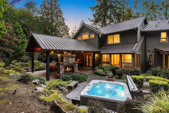 back house at dusk with a hot tub, an outdoor stone fireplace, and a patio area