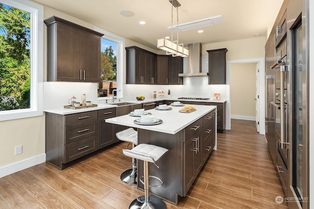 kitchen featuring wall chimney range hood, light countertops, dark brown cabinets, a kitchen island, and light wood finished floors