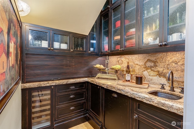 bar featuring dark brown cabinets, light stone counters, sink, beverage cooler, and backsplash