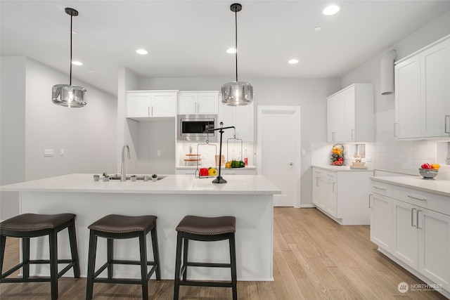 kitchen with stainless steel microwave, sink, decorative light fixtures, white cabinets, and an island with sink