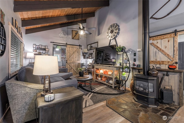 living room featuring ceiling fan, beam ceiling, a wood stove, hardwood / wood-style flooring, and a barn door