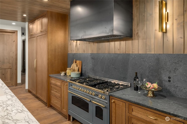 kitchen with dark stone counters, range with two ovens, light hardwood / wood-style flooring, wall chimney exhaust hood, and tasteful backsplash