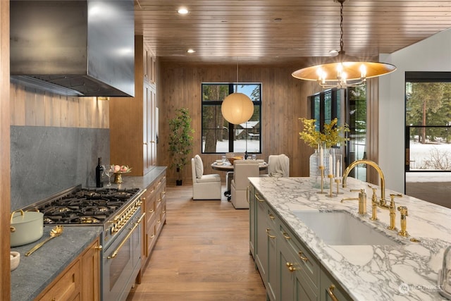 kitchen with light stone counters, stainless steel range, sink, wall chimney range hood, and hanging light fixtures