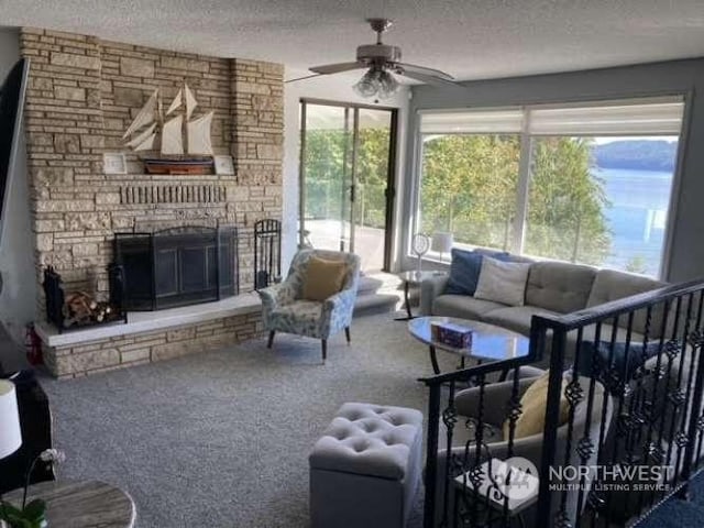 living room with ceiling fan, carpet floors, a stone fireplace, and a textured ceiling