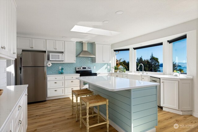 kitchen featuring appliances with stainless steel finishes, a skylight, light wood-type flooring, white cabinetry, and wall chimney exhaust hood