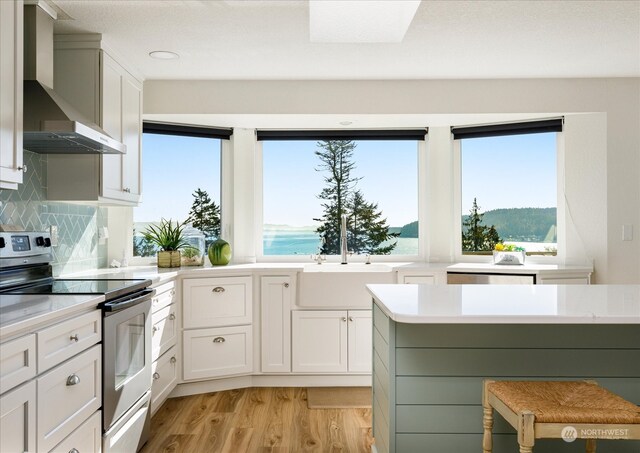 kitchen featuring wall chimney range hood, a water view, stainless steel electric range, and white cabinetry