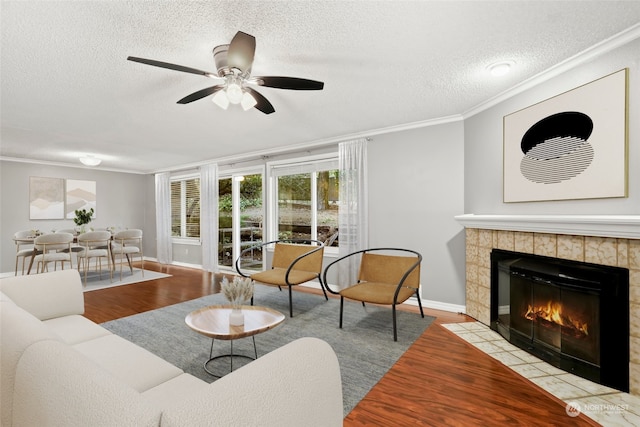 living room with ceiling fan, a textured ceiling, a tiled fireplace, crown molding, and light hardwood / wood-style floors