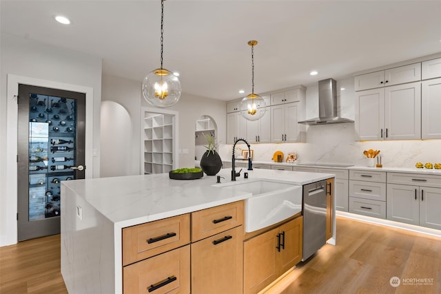 kitchen with wall chimney range hood, an island with sink, white cabinetry, stainless steel dishwasher, and light hardwood / wood-style flooring