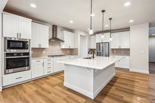 kitchen featuring wall chimney range hood, white cabinets, appliances with stainless steel finishes, light hardwood / wood-style flooring, and decorative light fixtures