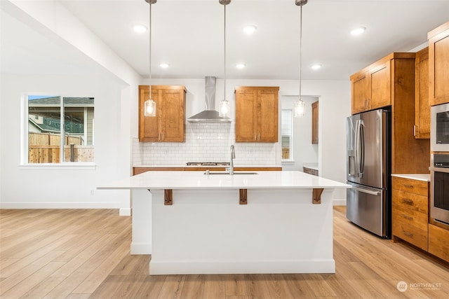 kitchen with a center island with sink, wall chimney range hood, sink, decorative light fixtures, and stainless steel appliances