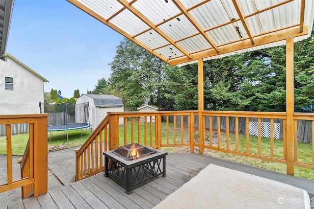 wooden terrace with a fire pit, a trampoline, a lawn, and a shed