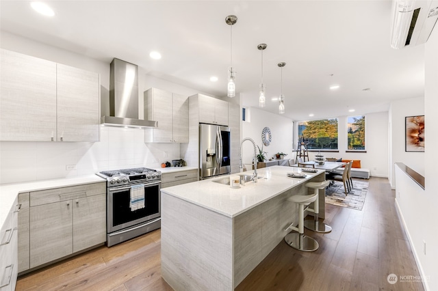 kitchen with sink, a center island with sink, wall chimney range hood, stainless steel appliances, and light hardwood / wood-style floors