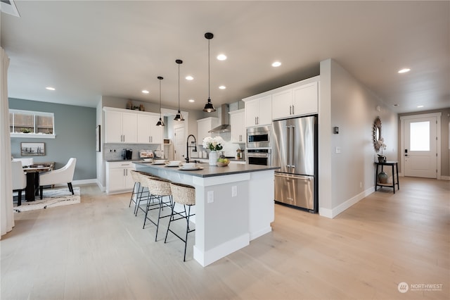 kitchen with wall chimney exhaust hood, light hardwood / wood-style floors, a center island with sink, appliances with stainless steel finishes, and white cabinetry