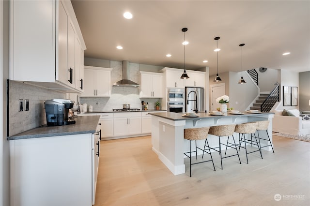 kitchen featuring a kitchen island with sink, stainless steel appliances, white cabinets, light hardwood / wood-style floors, and wall chimney range hood