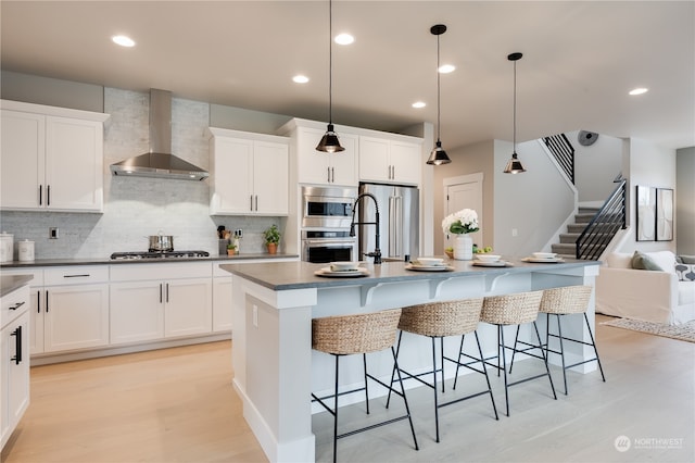 kitchen with white cabinets, an island with sink, hanging light fixtures, wall chimney range hood, and appliances with stainless steel finishes