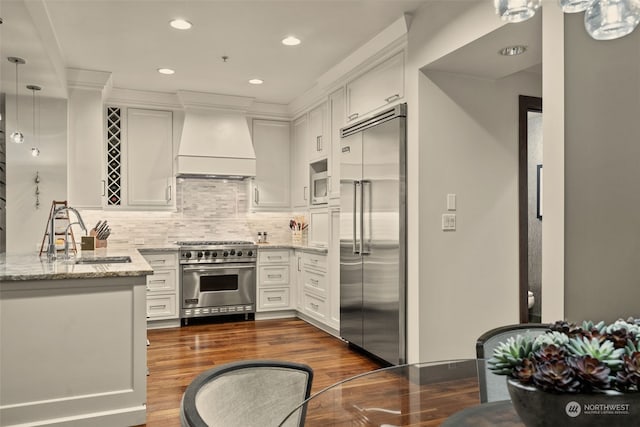 kitchen featuring sink, custom exhaust hood, light stone counters, built in appliances, and decorative light fixtures