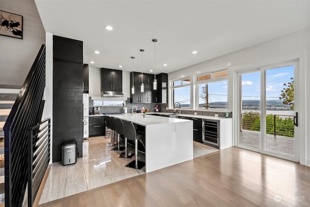 kitchen with a kitchen island with sink, beverage cooler, light countertops, and dark cabinetry