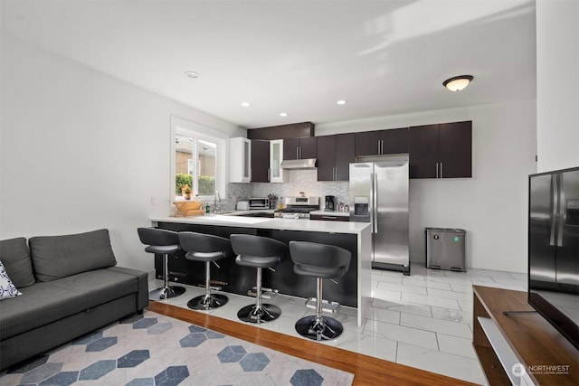 kitchen featuring a peninsula, a breakfast bar, under cabinet range hood, light countertops, and stainless steel appliances