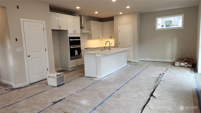 kitchen featuring a center island with sink, white cabinets, sink, double wall oven, and decorative backsplash